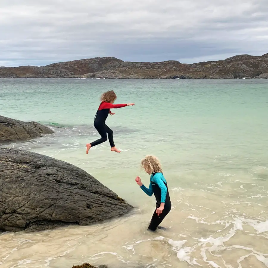 2 children in wetsuits in the Scottish sea, one walking through the water and the other jumping off a rock into the water wearing 2barefeet wetsuits