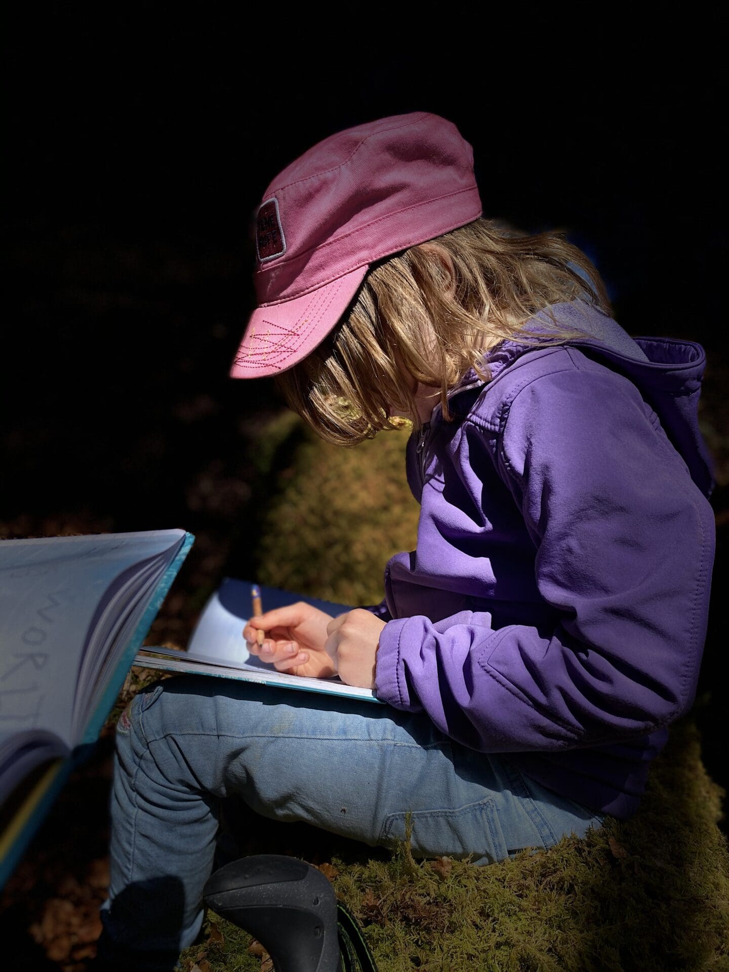 child drawing in a nature journal wearing pink cap and purple jumper