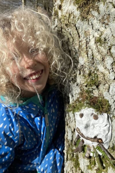 child with curly hair and blue Muddy Puddles jacket standing beside a tree with a clay face and natural materials used as hair and eyes