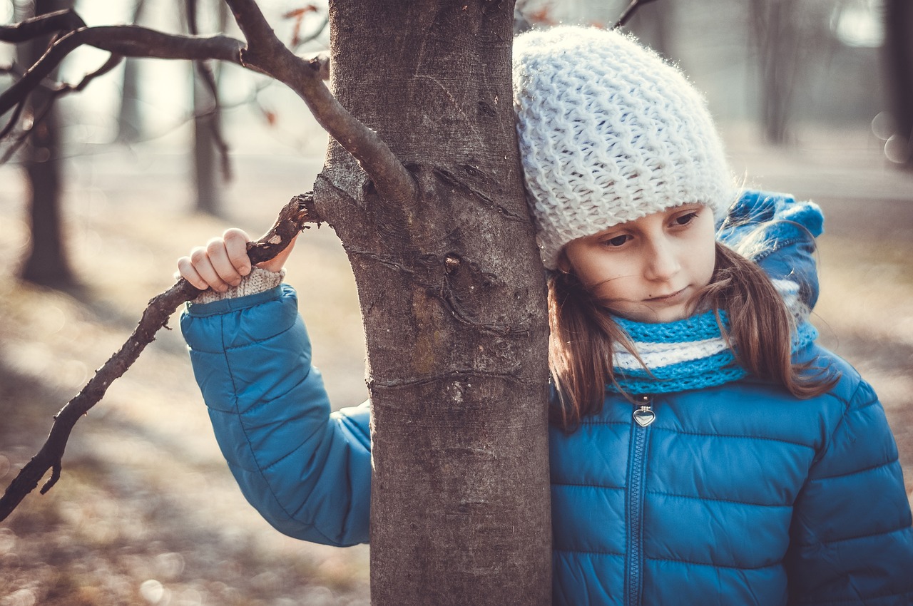 Child looking sad holding onto a tree with winter clothing